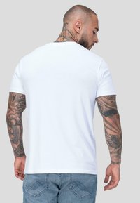 Kaldpea mehine, kellel on täielikud käetätoveeringud, kannab lihtsat valget T-särki ja siniseid teksapükse, seistes näoga ära heledashallil taustal.