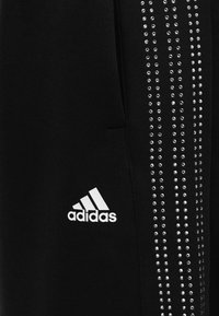 Μαύρο αθλητικό παντελόνι με ασημένιες λεπτομέρειες με πλινθώματα κατά μήκος της πλευράς, με λευκό λογότυπο Adidas στο κάτω αριστερό μέρος. Απαλή υφή υφάσματος.