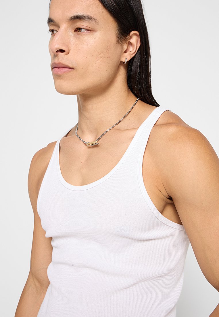 T-shirt sans manches blanc en tissu côtelé, avec un col ras du cou, porté avec un collier en chaîne argenté et un simple accent doré.