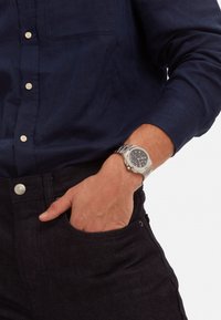 Montre-bracelet en acier inoxydable avec un cadran sombre, des fonctionnalités de chronographe et un bracelet en argent, portée sur une main dans une poche de pantalon en denim foncé.