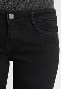 Jeans de mezclilla negra con un diseño ajustado, estilo de cinco bolsillos y cierre con botón de metal. La tela tiene una textura suave con detalles de costura sutiles.