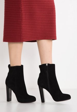 Bottines en daim noir, à bout rond, avec un talon haut et une fermeture éclair sur le côté. Le design présente un bord supérieur froncé pour une texture supplémentaire.