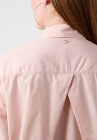 Kvinna i en ljusrosa skjorta med en bakfåll och en liten metallisk logotyp nära kragen, samt stora silverfärgade hoop-örhängen.