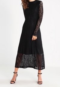 Kvinna som bär en svart spets långärmad midiklänning med genomskinlig nederkant och svarta öppna tå ankelsandaler, stående mot en vit bakgrund.