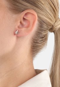 Primo piano dell'orecchio di una donna bionda che indossa un piccolo orecchino a forma di foglia in argento con pietre trasparenti incastonate.