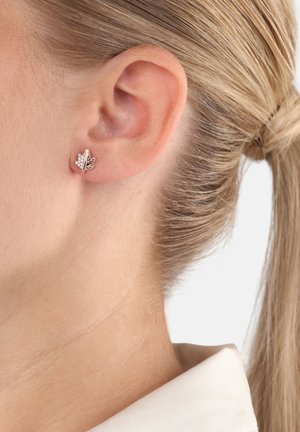 Primer plano de la oreja de una mujer rubia que lleva un pequeño pendiente en forma de hoja de plata con piedras transparentes incrustadas.