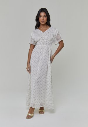 Witte maxi-jurk met polkadots, korte klokmouwen, een V-hals en een getailleerde taille. Gecombineerd met gouden slip-on sandalen. Glad, vloeiend materiaal.
