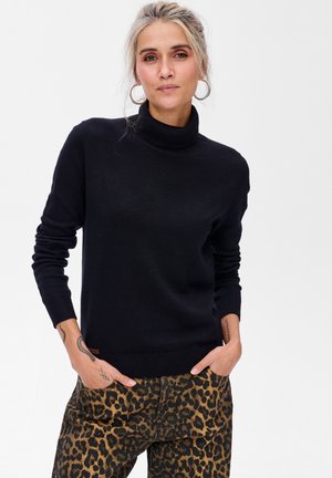 Kobieta z blond włosami w czarnej bluzie z golfem i spodniami w panterkę, z rękami w kieszeniach, stojąca na białym tle.