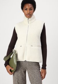 Krämfärgad fleeceväst med hög krage och framzip, med sidofickor med dragkedjor. Bärs över en brun långärmad topp.