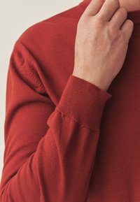 Pull rouge en tricot avec une coupe ajustée, doté d'un poignet texturé et de détails subtils de couture. Le matériau semble doux et léger.