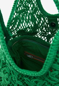 Grön vävd tote-väska med ett nätliknande mönster. Inuti finns en ficka med dragkedja med bruna detaljer och en mjuk grön foder.