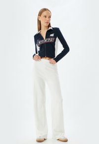 Giacca corta blu navy con zip e testo "LEGENDS", pannelli laterali bianchi. Paio di pantaloni bianchi a gamba larga con una texture liscia e una vita alta.