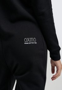 Čierna mikina so priliehavým strihom, ribbovanými manžetami a vyšitým logom s nápisom "OXMO" na zadnej vrecku, spolu s textom nižšie.