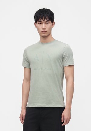 Junger Mann mit kurzen schwarzen Haaren, der ein hellgrünes Armani Exchange T-Shirt und schwarze Hose trägt, vor einem schlichten weißen Hintergrund stehend.