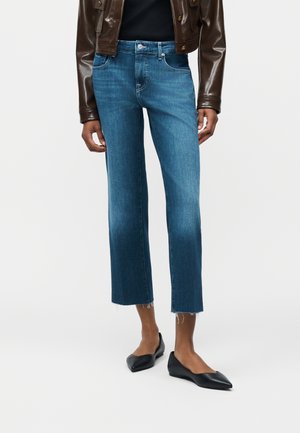 Femme portant un jean court bleu avec un ourlet brut, des ballerines noires pointues, un haut noir, et une veste courte en cuir marron.