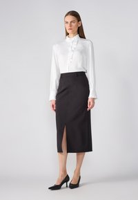 Blouse blanche avec un col à volants et une fermeture à boutons, associée à une jupe crayon noire avec une fente sur le devant, portée avec des talons noirs.