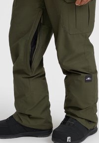 Olivgrüne Schneehosen mit seitlichen Reißverschlüssen, verstärkten Nähten und mehreren Taschen. Kombiniert mit schwarzen Schneestiefeln mit einem sicheren Riemen.