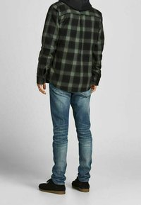 Camicia di flanella a quadri neri e verdi con maniche lunghe, abbinata a jeans blu sbiaditi e scarpe nere. Mostrata da dietro.
