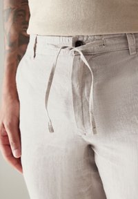 Pantaloni in lino grigi chiari con una cintura con cordino, caratterizzati da una trama liscia e un dettaglio a bottone sul davanti. Vista ravvicinata.