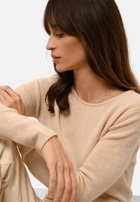 Beige gestrickter Pullover mit langen Ärmeln, weicher Textur und runder Ausschnitt; das Modell sitzt und zeigt eine angepasste Haltung und Frisur.
