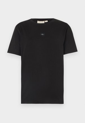 Czarna koszulka z krótkim rękawem i okrągłym dekoltem, z małym logo Calvin Klein na środku klatki piersiowej, przedstawiona na jasnym tle.