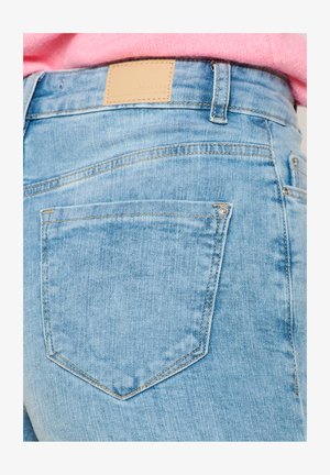 Light blue denim jeans feature a smooth texture, five-pocket design, and a tan leather tag at the waistband with embossed logo detail.
