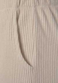 Beige geripptes Material mit einer Seitentasche und einem elastischen Bund, das eine weiche Textur mit subtilen horizontalen Linien vermittelt.