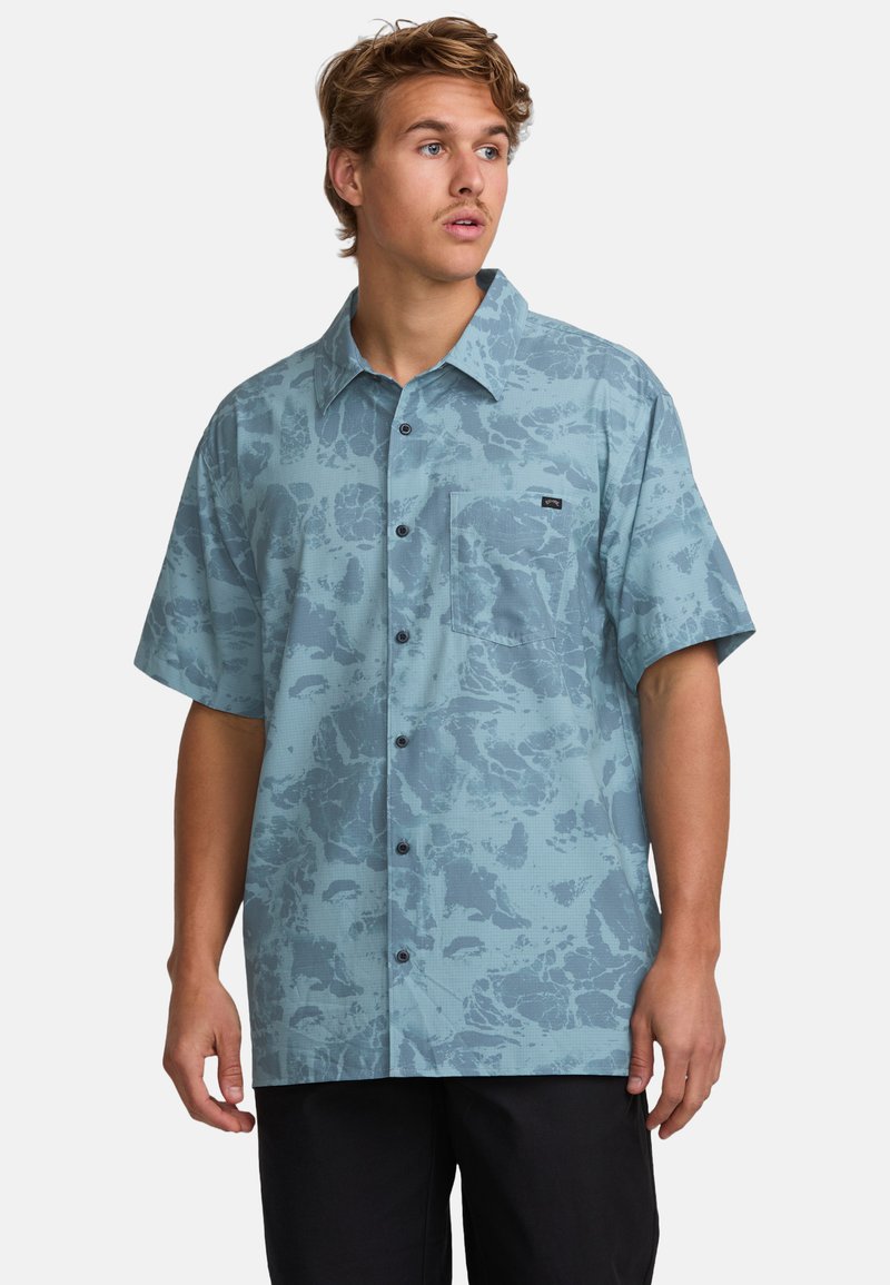 Young man wearing a short-sleeve blue patterned button-up shirt with a chest pocket, looking to the side against a plain background.