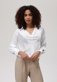 Blusa blanca de seda con un escote drapeado, mangas largas que presentan acentos de botones, combinada con pantalones beige de corte holgado.
