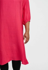 Luftige, pinkfarbene Tunika aus strukturiertem Stoff, mit Dreiviertelärmeln und asymmetrischem Saum, kombiniert mit schwarzen Leggings.