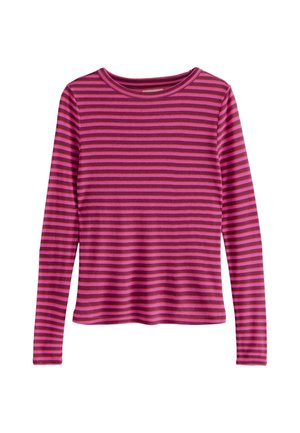 Ung kvinde med langt mørkt hår iført en rød og mørk pink stribet langærmet trøje og sorte bukser, står foran en ensfarvet grå baggrund.