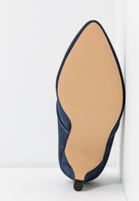 Chaussure à talon haut en daim bleu marine avec un bout pointu, une tige texturée et une semelle lisse couleur camel. Présente un talon étroit pour une silhouette élégante.