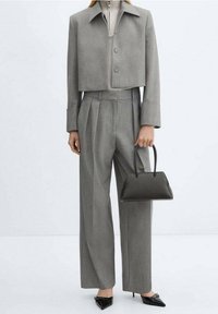 Blazer gris corto con cuello puntiagudo, combinado con pantalones grises plisados de tiro alto. La modelo sostiene un pequeño bolso negro y lleva tacones negros puntiagudos.