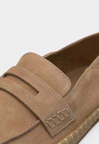 Mocassin en daim marron clair avec une large lanière traversant le dessus et une semelle en jute tressé, affichant des coutures détaillées sur la lanière et la tige.