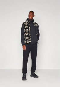 Juodas fliso liemenė su baltais geometriniais raštais, dėvima ant juodo palaidinės, derinama su juodomis kelnėmis ir juodais sportbačiais.