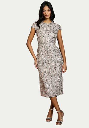 Donna che indossa un vestito midi aderente decorato con paillettes argentate, con maniche corte e sandali con tacco alto e cinturino alla caviglia, in piedi con una gamba incrociata.
