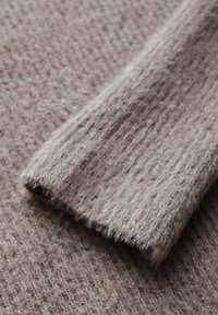 Ljusbrun stickad tyg med en mjuk textur och ett ribbat mönster. Kanterna är något fransiga, vilket ger ett avslappnat inslag till materialet.