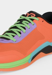 Πορτοκαλί αθλητικό παπούτσι με πράσινα κορδόνια, μοβ πάνελ με οπές για κορδόνια, μαύρη γλώσσα και πολύχρωμη σόλα που περιλαμβάνει πράσινες, μπλε και μαύρες λεπτομέρειες.