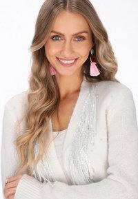 Fehér kötött kardigán gyöngy díszítéssel és ezüst flitteres bojtokkal, fehér felső fölött viselve. Rózsaszín bogyós fülbevalók színkontrasztot adnak.