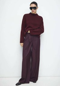 Maglione lavorato a maglia color bordeaux con collo alto abbinato a pantaloni a gamba larga color prugna intenso e occhiali da sole oversize neri. Texture lisce in tutto l'insieme.