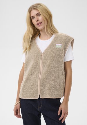 Kvinde med langt blondt hår bærer en beige struktureret fleecevest med lynlås over en hvid T-shirt og mørke jeans mod en simpel baggrund.