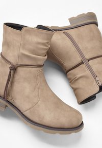 Stivali ankle in pelle sintetica beige con un design morbido, cerniera laterale e dettaglio decorativo in marrone a fiocco nella parte superiore. Suola in gomma spessa.