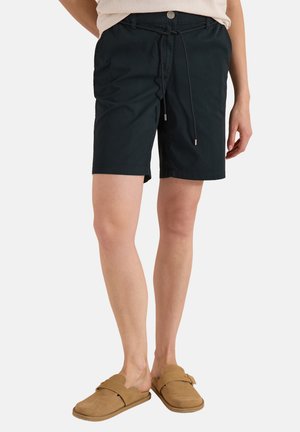 Sorte shorts med snøre, båret af en person, kombineret med lysebrune slip-on sko, stående mod en ensartet lys baggrund.