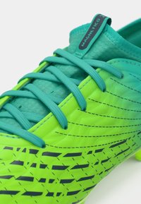 Green and turquoise soccer cleat with textured synthetic upper, prominent laces, and black geometric patterns. Includes a pull tab labeled "MAGNETICO."