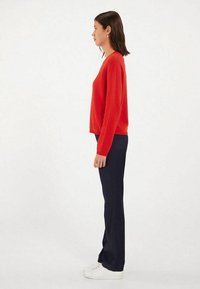 Pull rouge à manches longues, associé à un pantalon bleu marine et des baskets blanches. Présentant une coupe décontractée et un discret logo sur le pull.