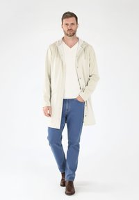 Chaqueta impermeable color beige claro con capucha, cierre de botones y bolsillos laterales, llevada sobre un suéter de pico color crema y jeans azules, combinada con botas marrones.