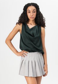 Top sin mangas de satén verde oscuro con escote drapeado, combinado con una falda corta plisada de color gris claro, que presenta una textura suave y plisados estructurados.