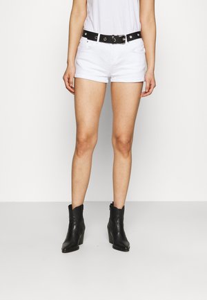 Person iført hvide shorts med sort bælte og sorte ankelstøvler står mod en ensartet lys baggrund.