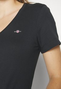 T-shirt noir à manches courtes avec col en V, avec un petit logo GANT rouge et blanc brodé sur la poitrine gauche, porté par une personne.