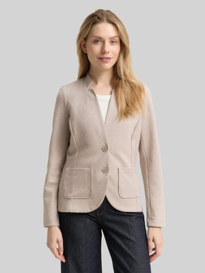 Blonde vrouw die een beige twee-knopen blazer met voorzakken draagt over een wit topje en donkere jeans, staand voor een effen grijze achtergrond.